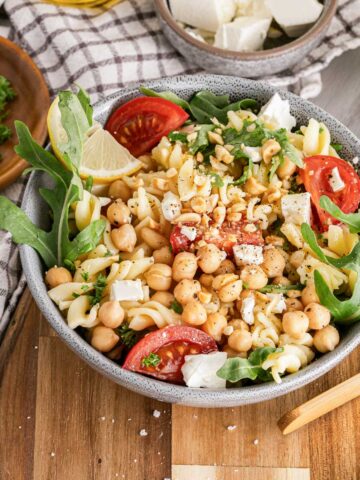 A bowl of chickpea salad with tomatoes, cheese, and arugula on a wooden table, garnished with herbs and lemon.