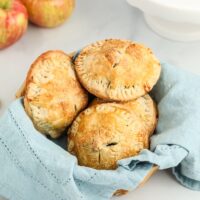 A basket lined with a blue cloth holds four golden-brown hand pies, with apples in the background.