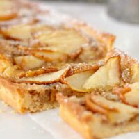 Close-up of a rectangular pear tart, dusted with powdered sugar, featuring thin slices of baked apple on a flaky pastry base, placed on a white surface.