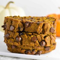 Three slices of pumpkin bread with chocolate chips, stacked on a white surface with blurred pumpkins in the background.