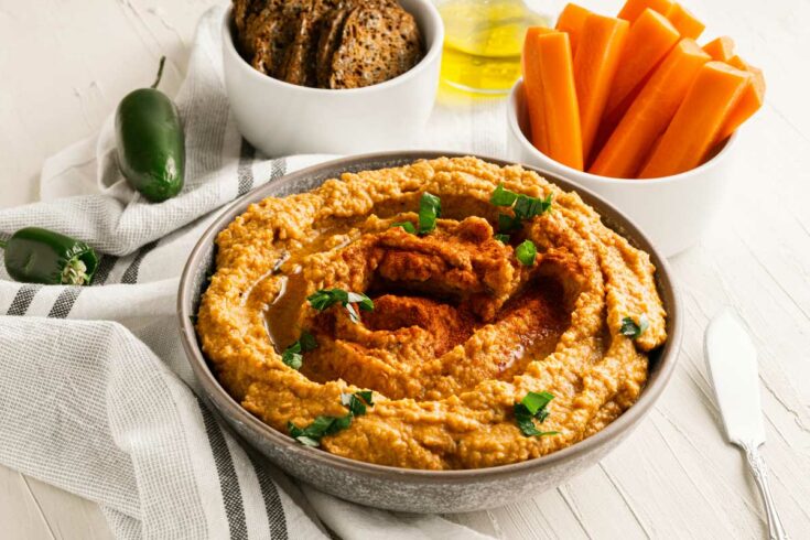 Bowl of roasted red pepper hummus with chipotle with bread crisps and carrot sticks.