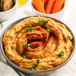 a bowl of Red pepper hummus with bread crackers and carrot sticks.