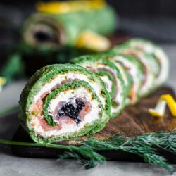 Close-up of sliced spinach roulade with salmon, cream cheese, and caviar filling, garnished with dill and lemon wedges, placed on a wooden board.