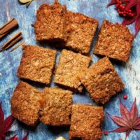 Nine oatmeal squares arranged on a rustic blue surface, surrounded by cinnamon sticks, apple slices, and red leaves.