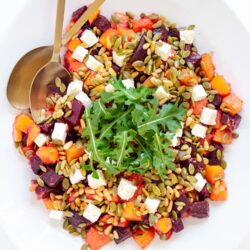 A fall salad with diced vegetables, feta cheese, pumpkin seeds, and a garnish of arugula, with a spoon and fork on the side.