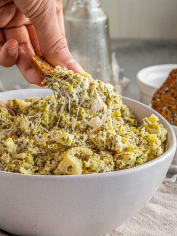 A hand dipping a cracker into a bowl of cheesy broccoli dip, with additional crackers in a dish in the background.