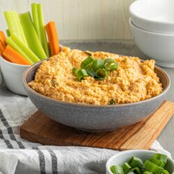 A bowl of buffalo cauliflower dip garnished with sliced green onions, placed on a wooden board, with celery and carrot sticks in a cup nearby and additional green onions in a small dish.
