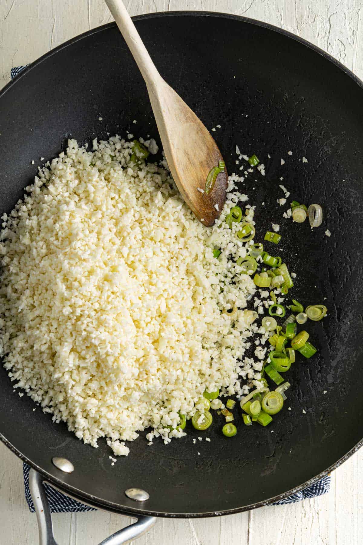 A black skillet with sautéed cauliflower rice and chopped green onions, stirred with a wooden spoon.