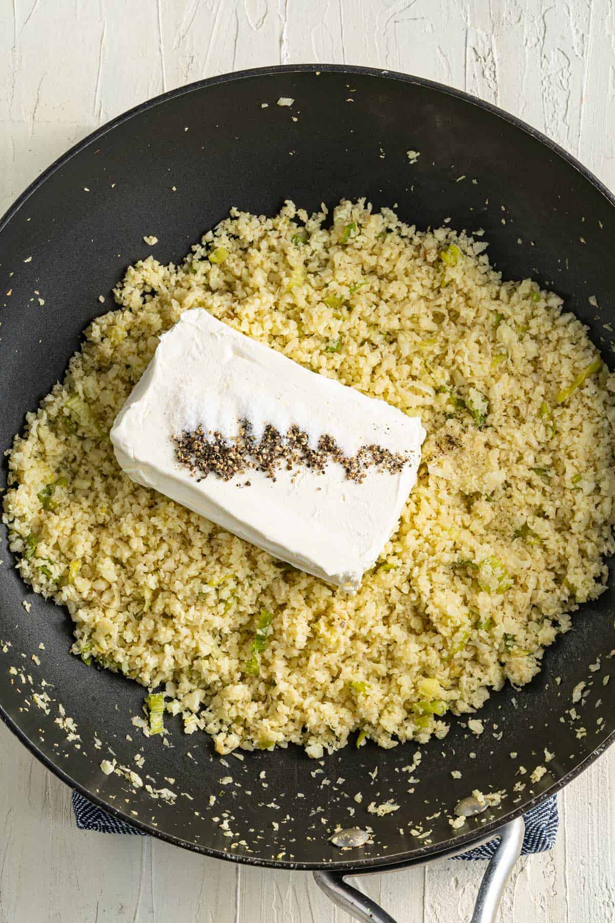 A large black skillet contains sautéed cauliflower rice with a block of cream cheese and a line of ground black pepper in the center.