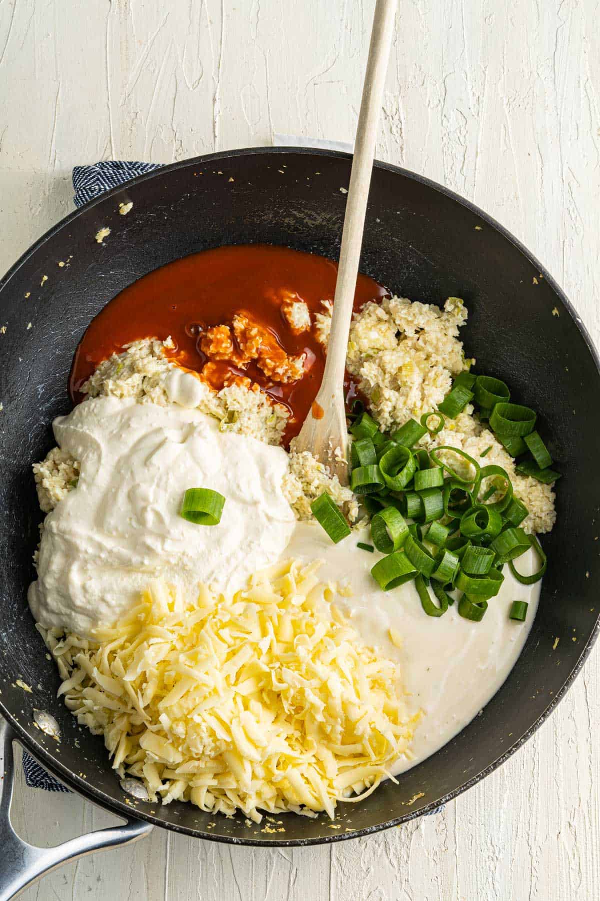 A skillet filled with buffalo sauce, cream, grated cheese, minced cauliflower, and chopped green onions, with a wooden spoon resting inside.
