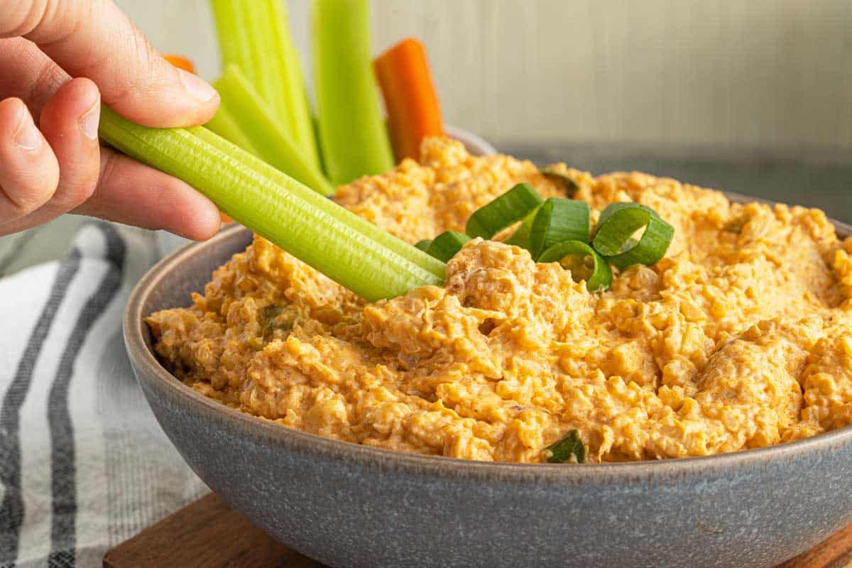 A hand dips a celery stick into a cauliflower buffalo dip topped with chopped green onions, with other vegetable sticks visible in the background.