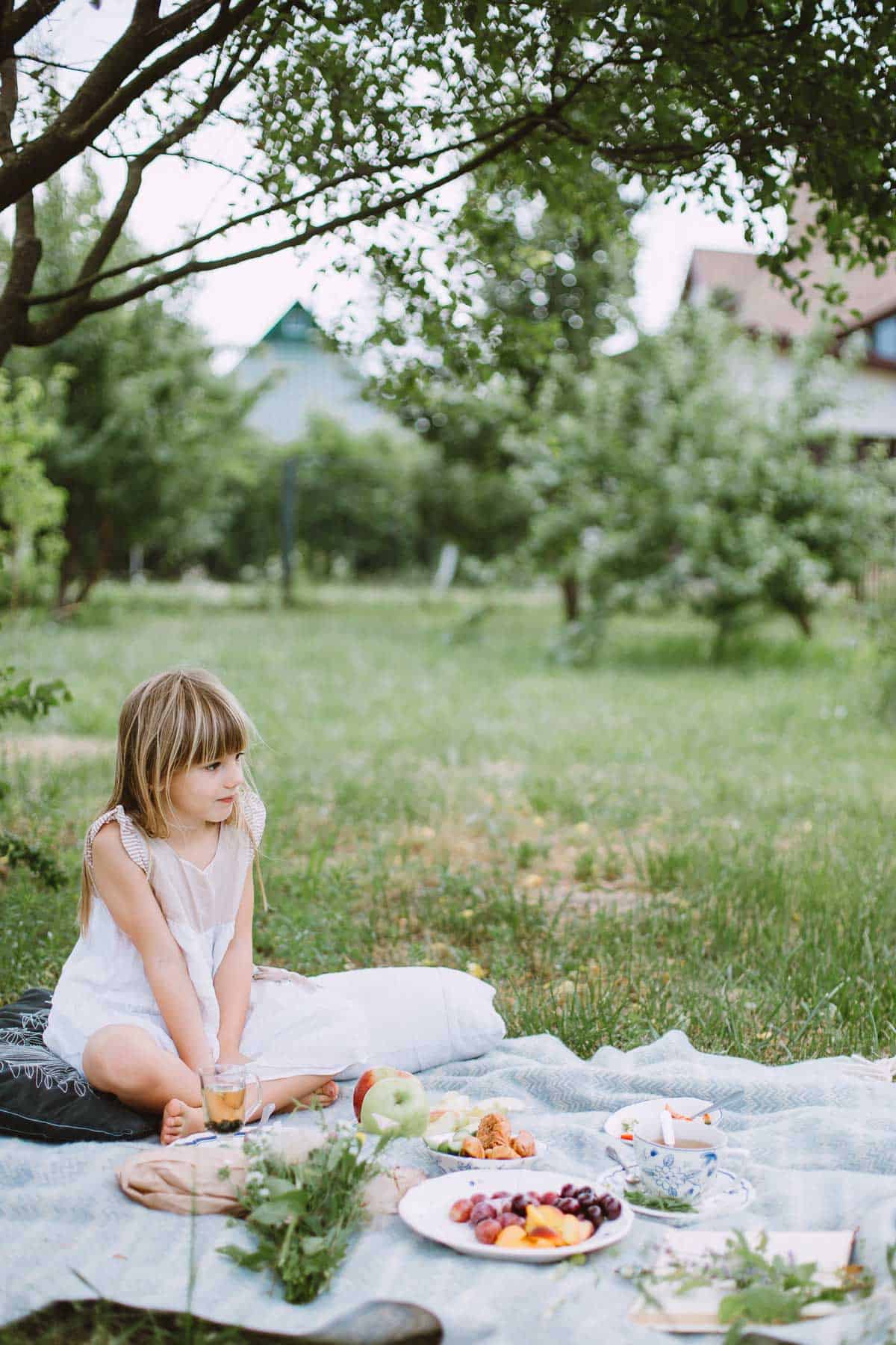 A little girl sitting on a blanket eating a picnic.