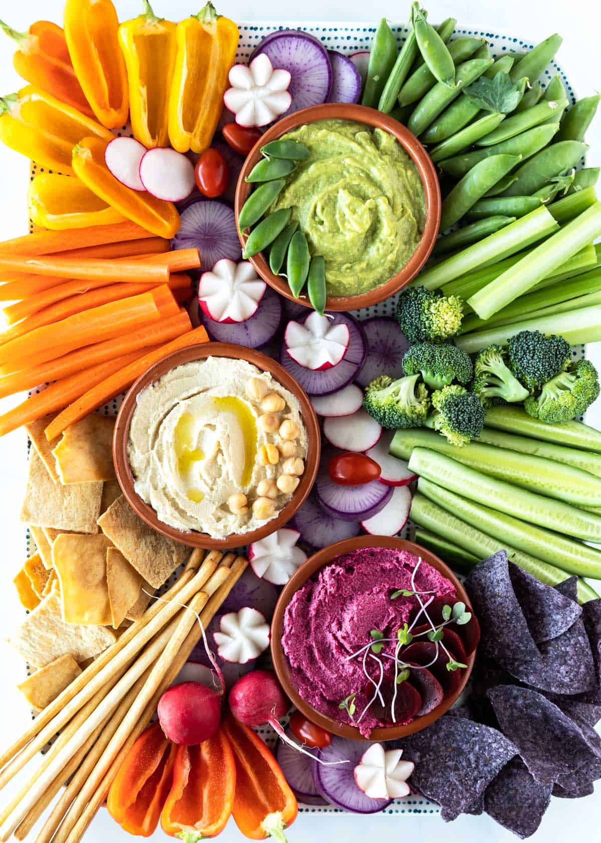 An assorted vegetable platter with three dips: green guacamole, white hummus, and purple beetroot hummus.