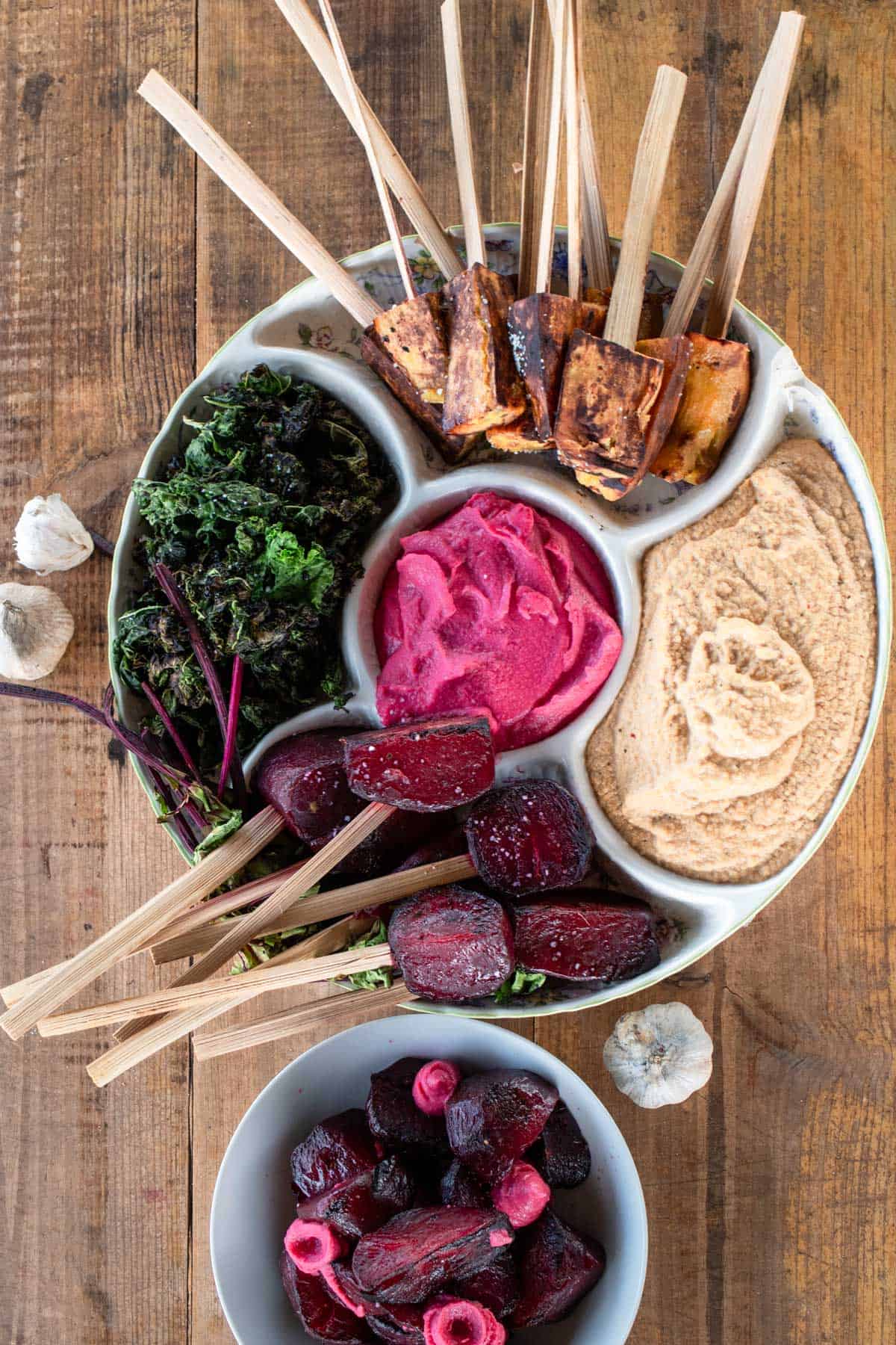 A round platter with skewered roasted vegetables, kale, beet hummus, traditional hummus, and a bowl of pickled beets on a wooden table.