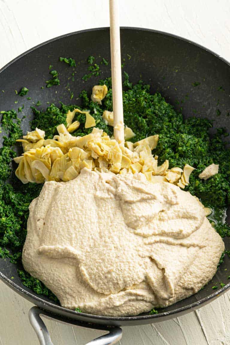 Chopped artichoke hearts, blended cashews and frozen spinach being mixed in a pan in preparation for a vegan dip.