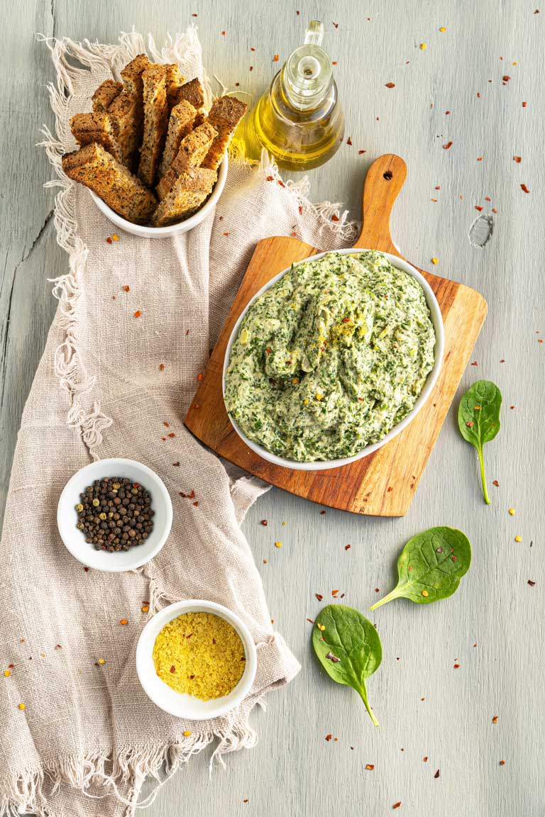 Vegan spinach dip on a board displayed with ingredients and dipping toasts.