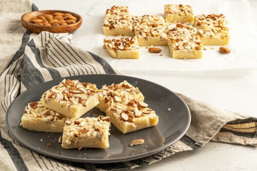 Squares of white chocolate fudge with almonds on a plate and on baking paper.