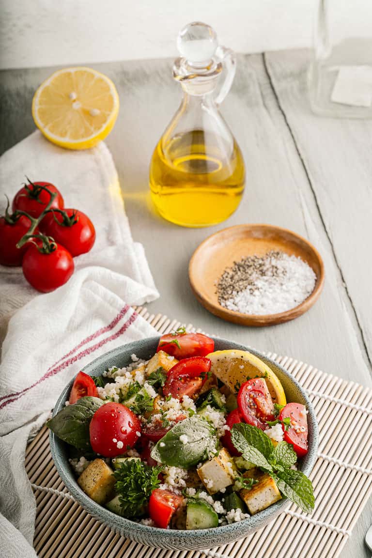 A bowl of couscous salad with tofu and fresh herbs on a table with olive oil.