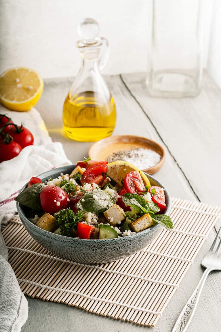 A bowl of couscous salad with tofu and fresh herbs on a table with olive oil. 