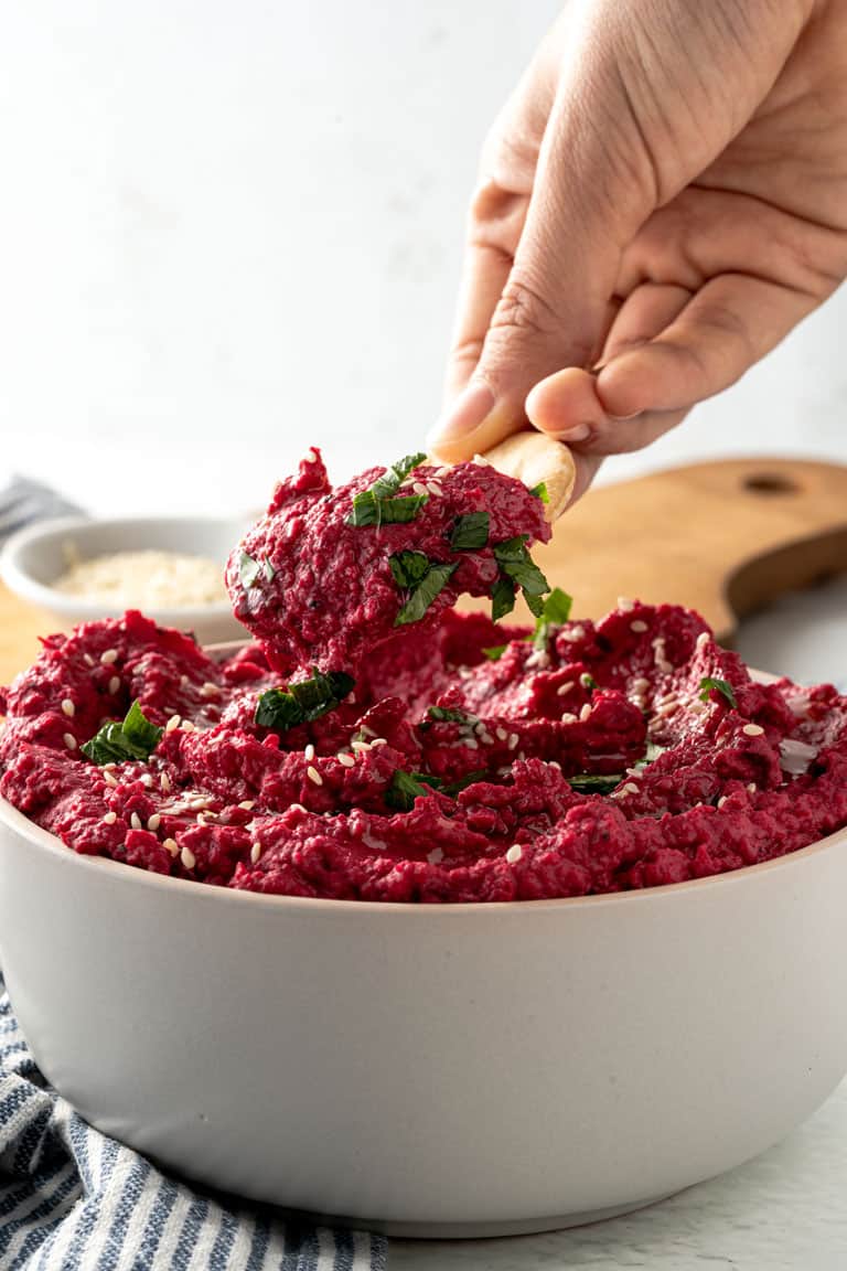 Dipping a cracker into a bright red bowl of healthy roasted beet hummus dip.