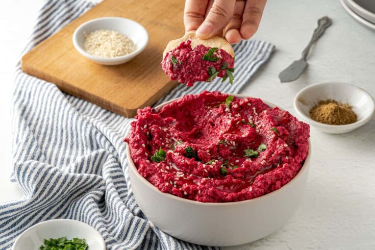 Dipping a cracker into a bright red bowl of healthy roasted beet hummus dip.