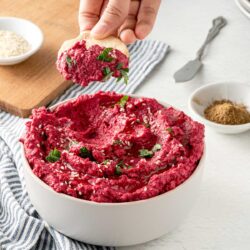 A vibrant bowl filled to the brim with creamy beet hummus, with a hand gently dipping into it.