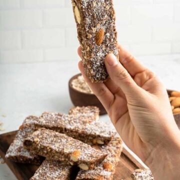 A hand holding a homemade granola bar with almonds and a sprinkle of coconut flakes, with more bars on a wooden board in the background.