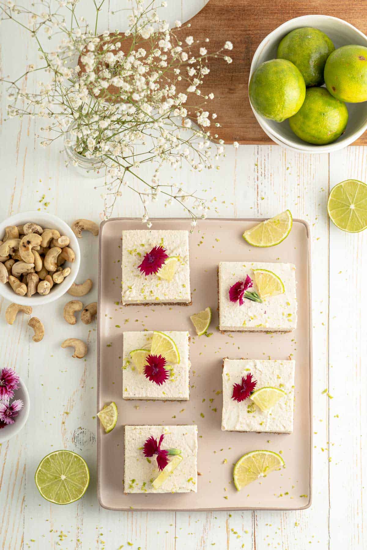 Key lime pie bars on a tray garnished with pink flowers surrounded by ingredients. 