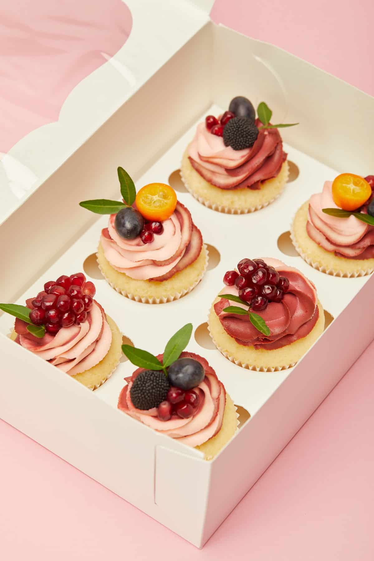 Cupcakes decorated with frosting and fruit in a cupcake box against a pink background. 