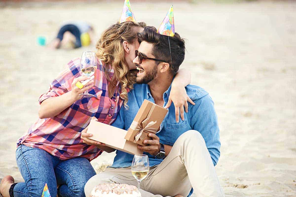 A couple celebrating a birthday on the beach. She is giving him a gift and a hug. 