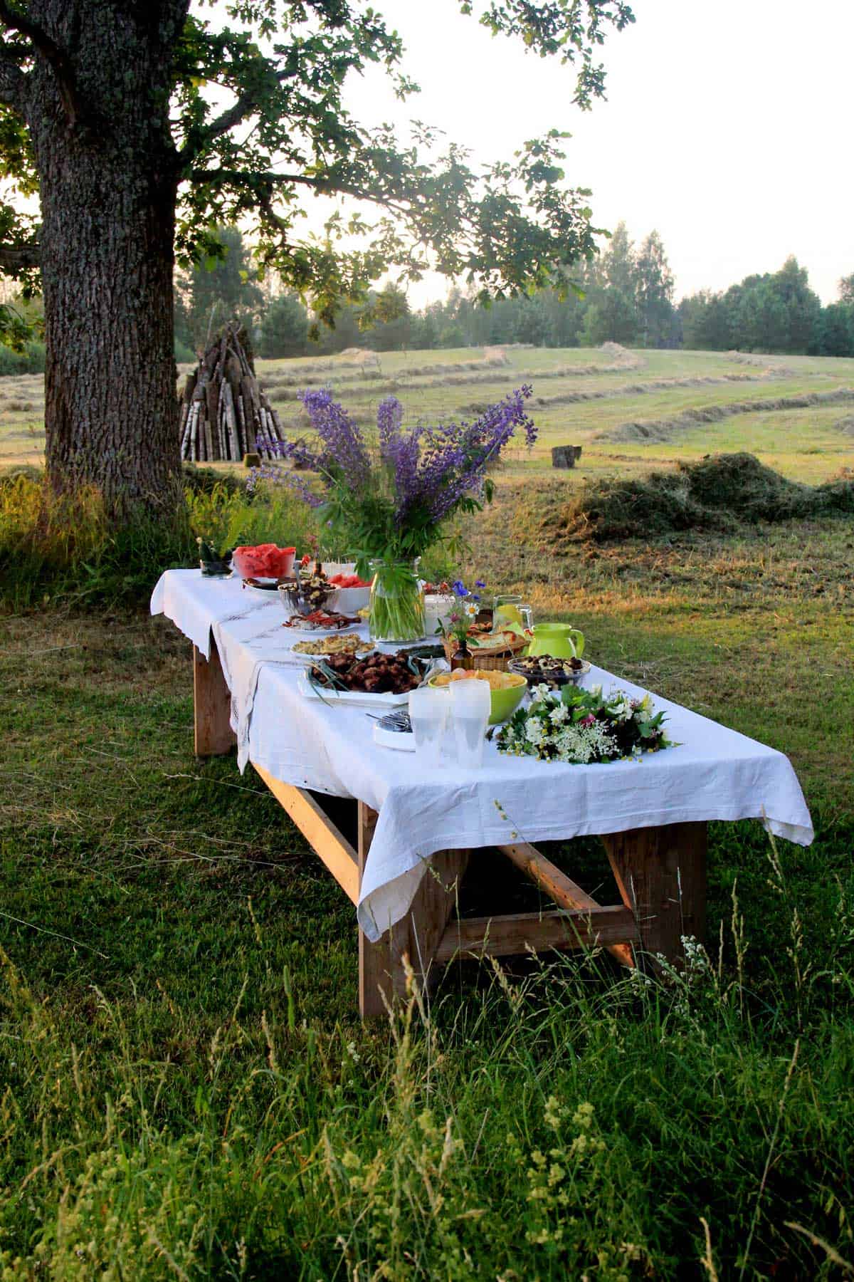 A picnic table in a field with a white table cloth and a large vase of purple flowers in the center.