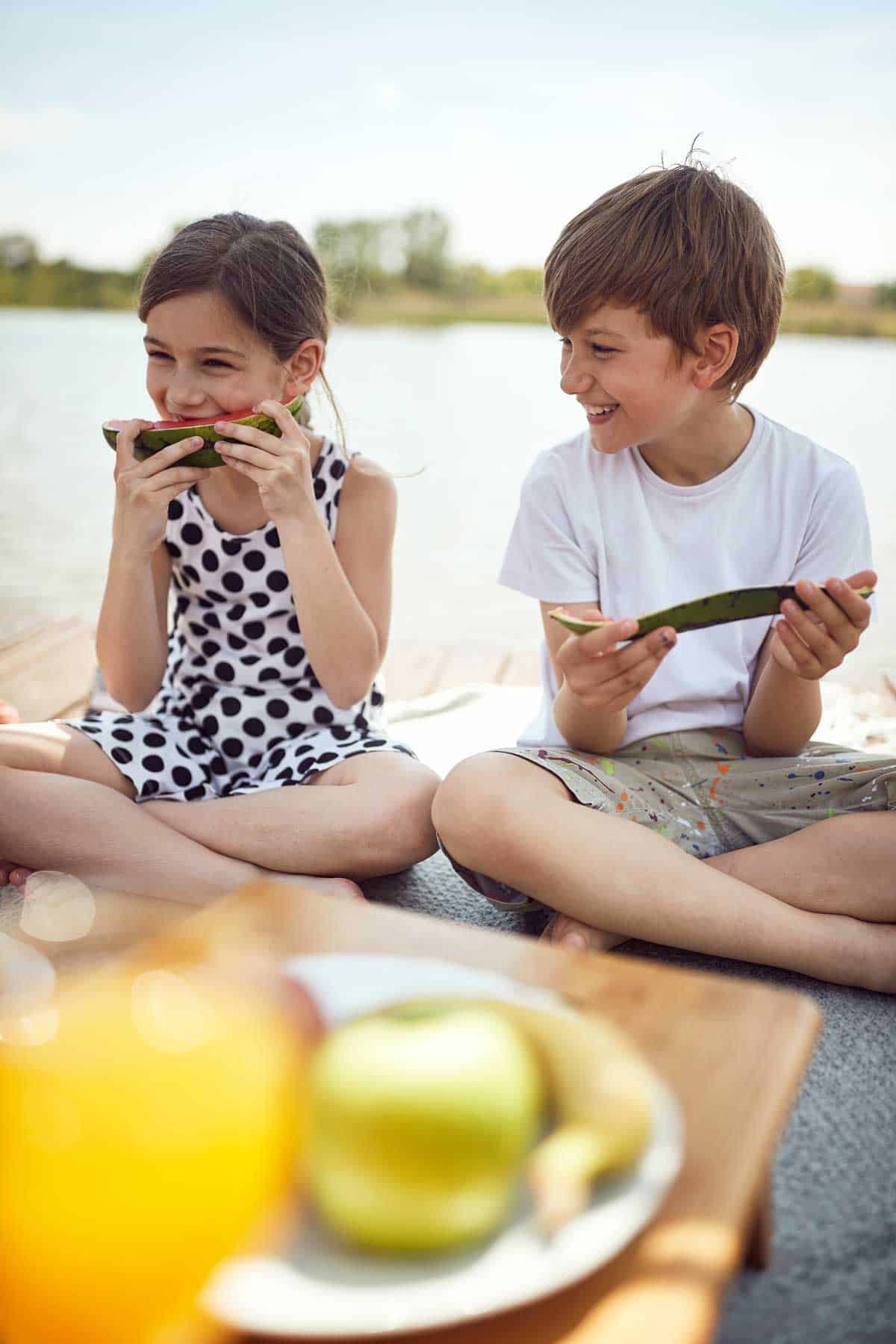 Two children sitting on the ground eating fruit at a picnic.