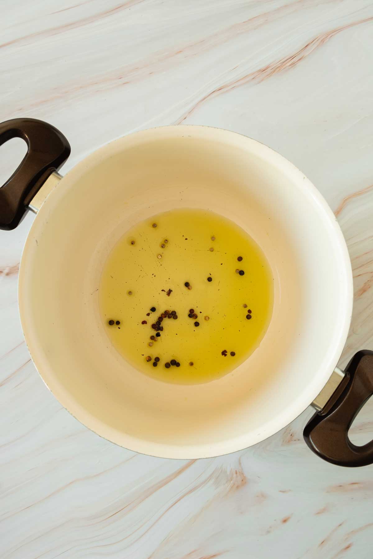 Olive oil in a pot with black pepper corns on a marble table.