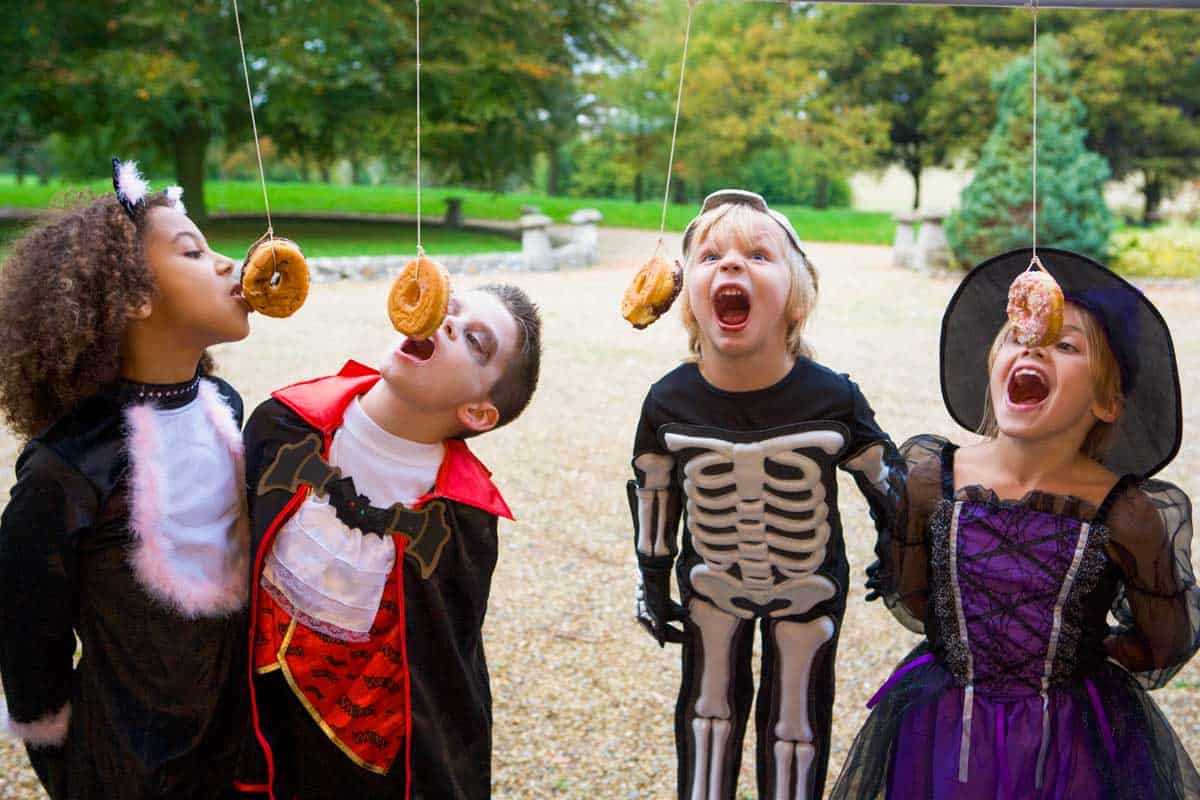A group of children dressed up in halloween costumes biting at hanging doughnuts.