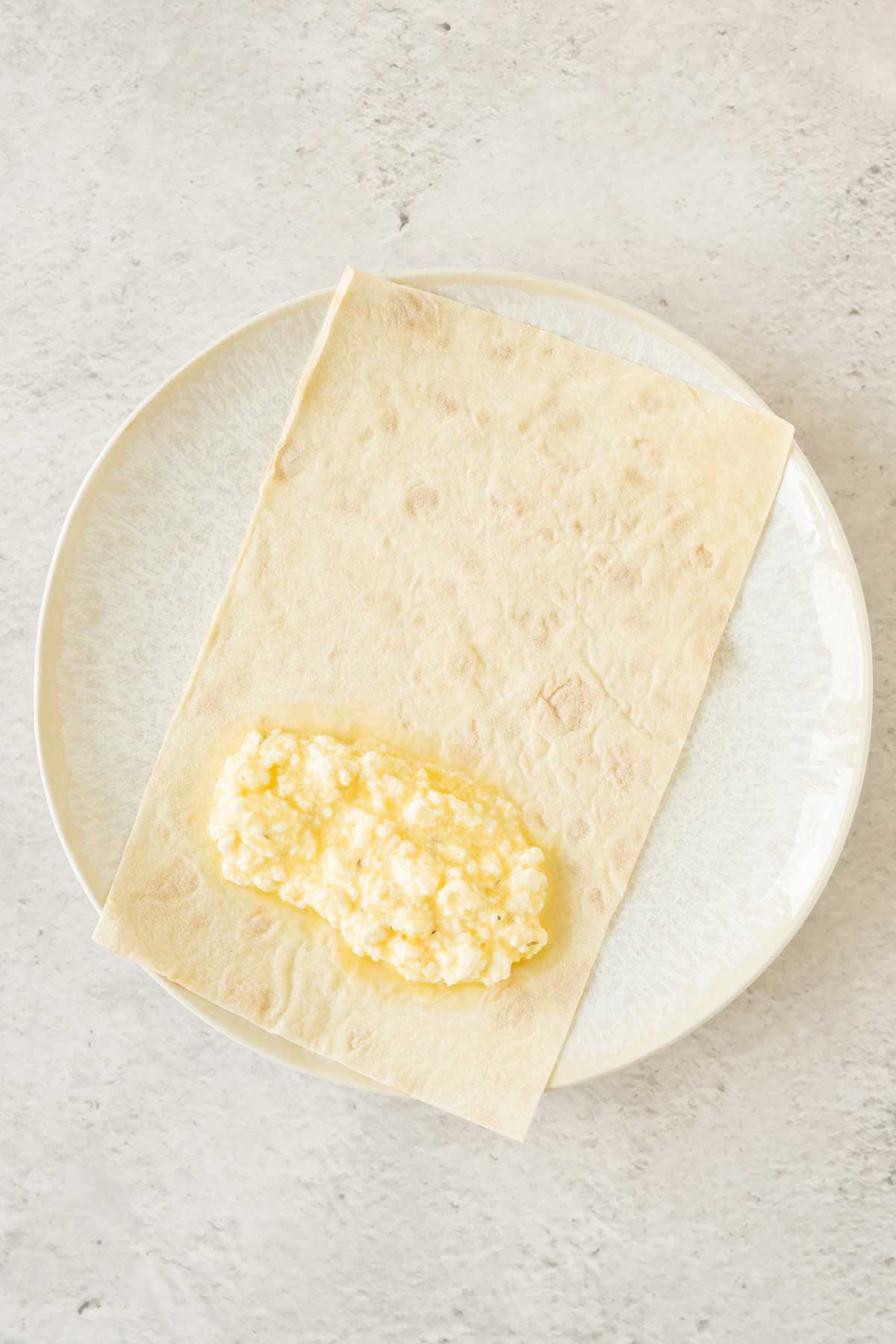 A white plate with a piece of filo pastry and the cheese and egg mix ready to be rolled.