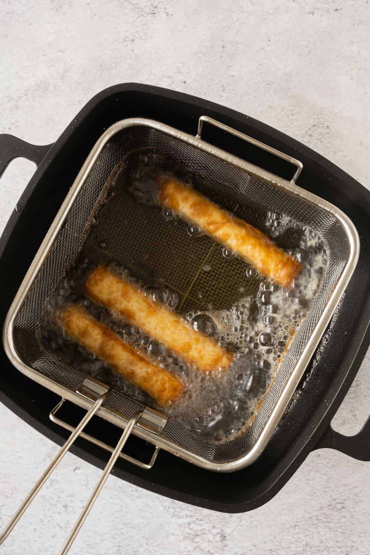 Cheese pastry rolls frying in a frying basket in oil.