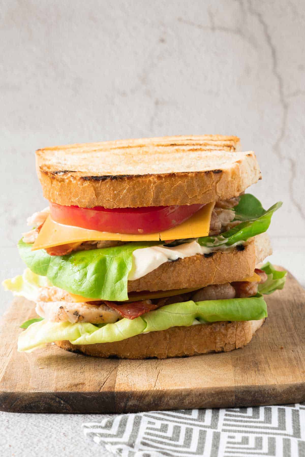 A layered grilled chicken club sandwich on a wooden cutting board.