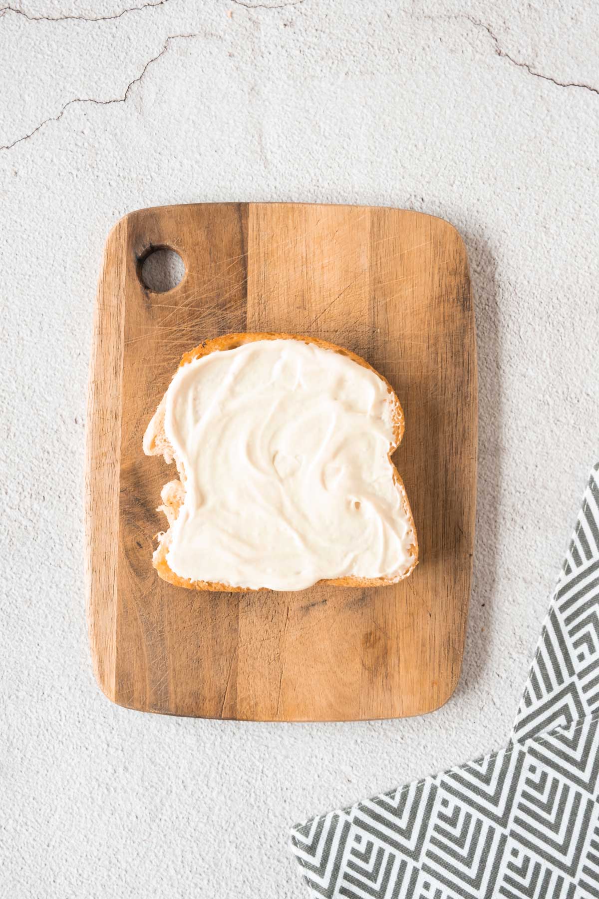 A slice of bread with mayonnaise on a wooden cutting board.