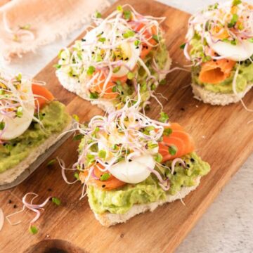 A wooden cutting board Avocado, smoked salmon, egg, and sprouts heart shaped sandwiches.
