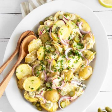 Dill pickle potato salad on a serving plate with wooden serving spoons on a white picnic table.