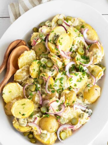 Dill pickle potato salad on a serving plate with wooden serving spoons on a white picnic table.