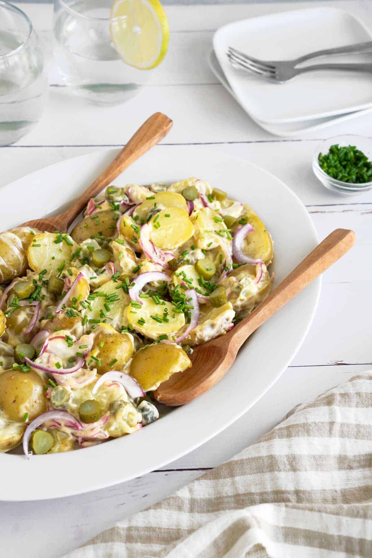 Dill pickle potato salad on a serving plate with wooden serving spoons on a white picnic table. 