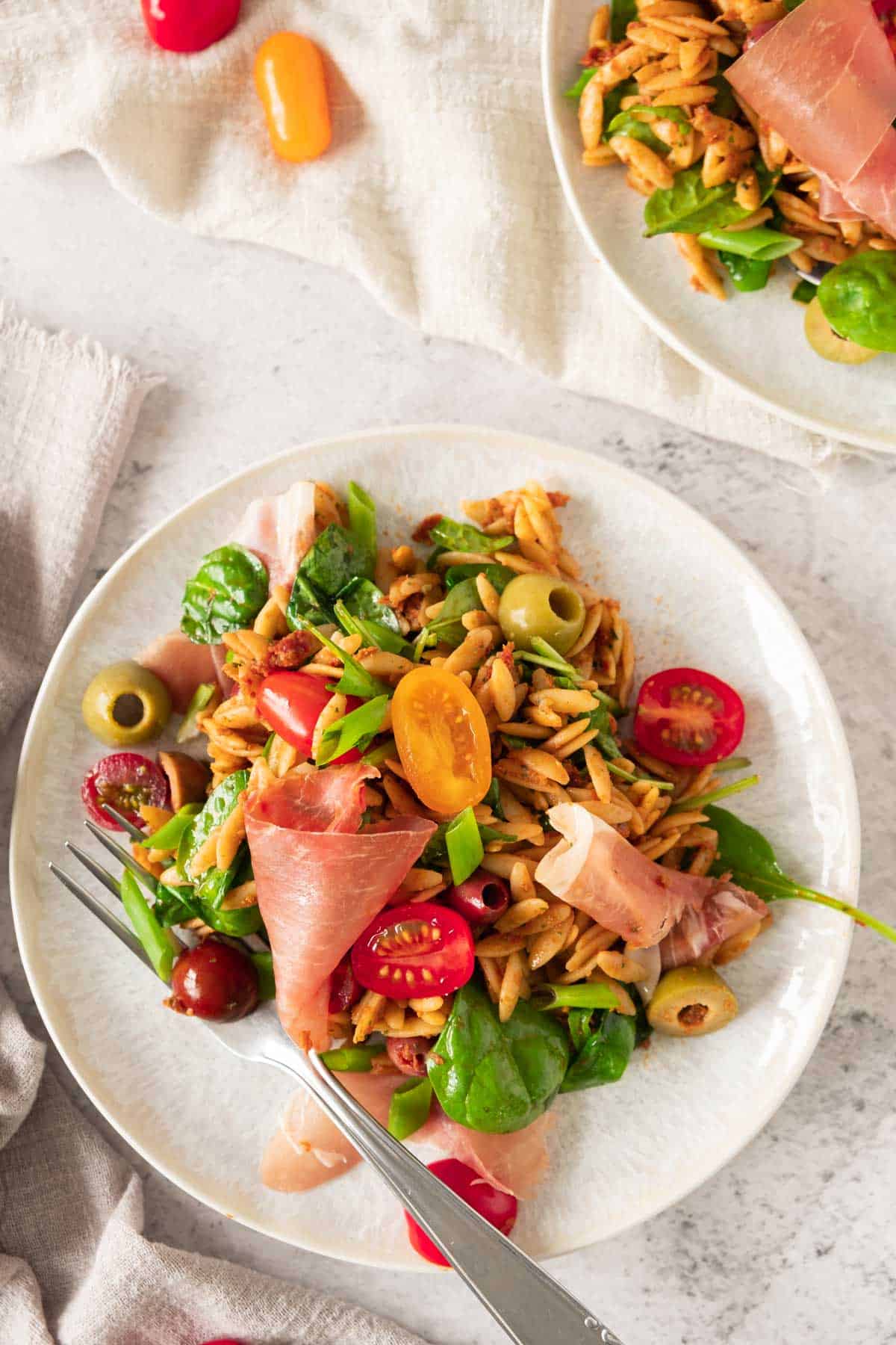 Pesto Orzo salad with parma ham, tomatoes and olives on a white plate.