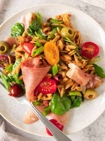 Pesto Orzo salad with parma ham, tomatoes and olives on a white plate.