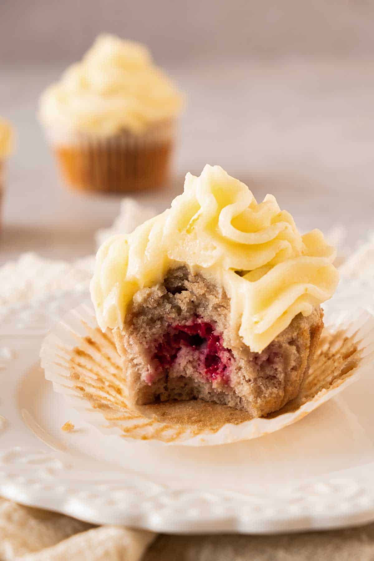 A raspberry cupcake with frosting on a cooling rack with a bite taken out of it.