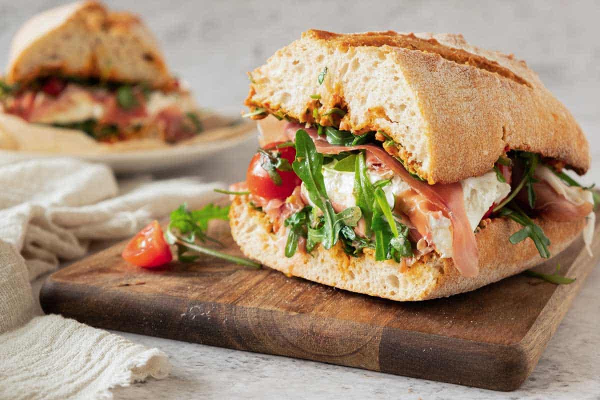 A fresh ciabatta sandwich with prosciutto, burrata, arugula, and tomatoes on a wooden cutting board.