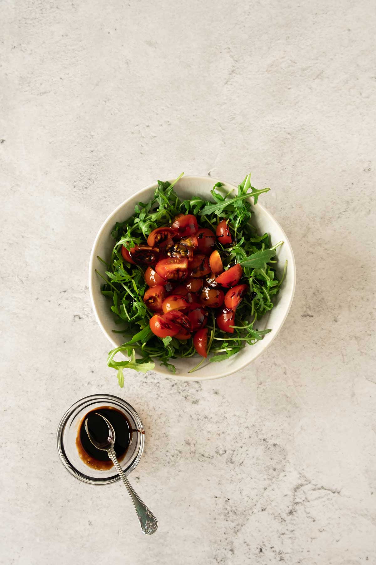 A bowl of arugula and cherry tomato salad with a side of balsamic dressing on a light surface.
