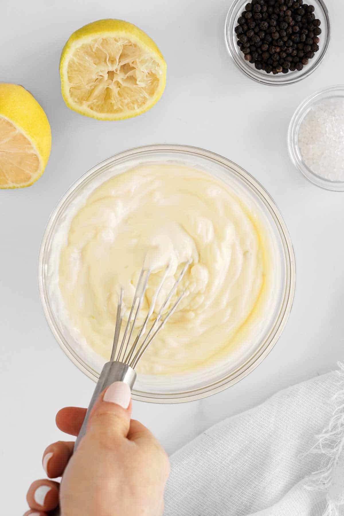 Whisking chicken salad dressing in a bowl with lemon and condiments on a white surface.