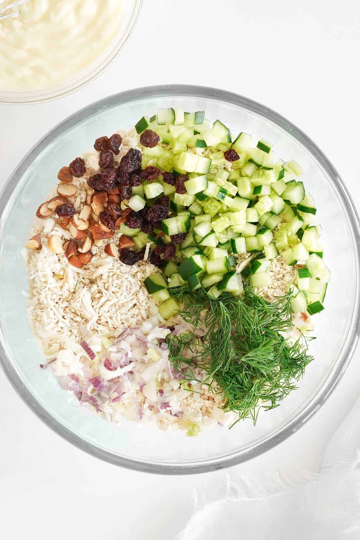 A glass bowl containing a mixture of chopped cucumber, shredded chicken, raisins, sliced almonds, chopped red onion, and fresh dill.