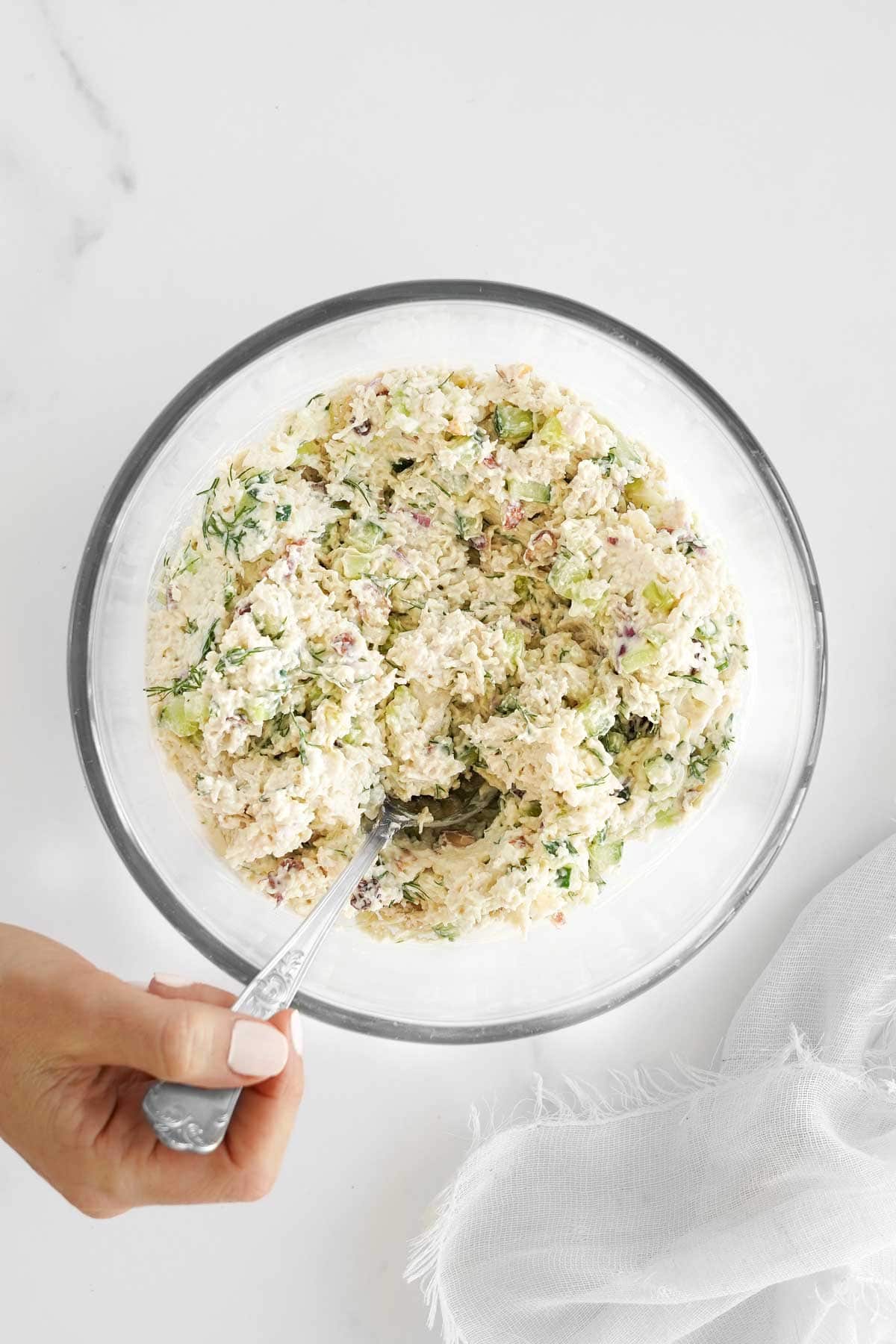 A bowl of creamy chicken salad with a hand mixing it using a spoon.